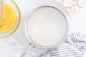 A sauce pan with cream sits next to eggs and a towel.