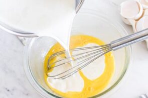 Pouring heavy cream into a bowl of egg yolks with a whisk inside the bowl.