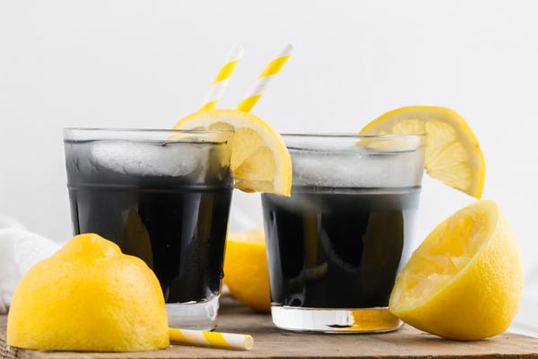 two small glasses filled with a black lemonade with striped yellow straws and lemons nearby