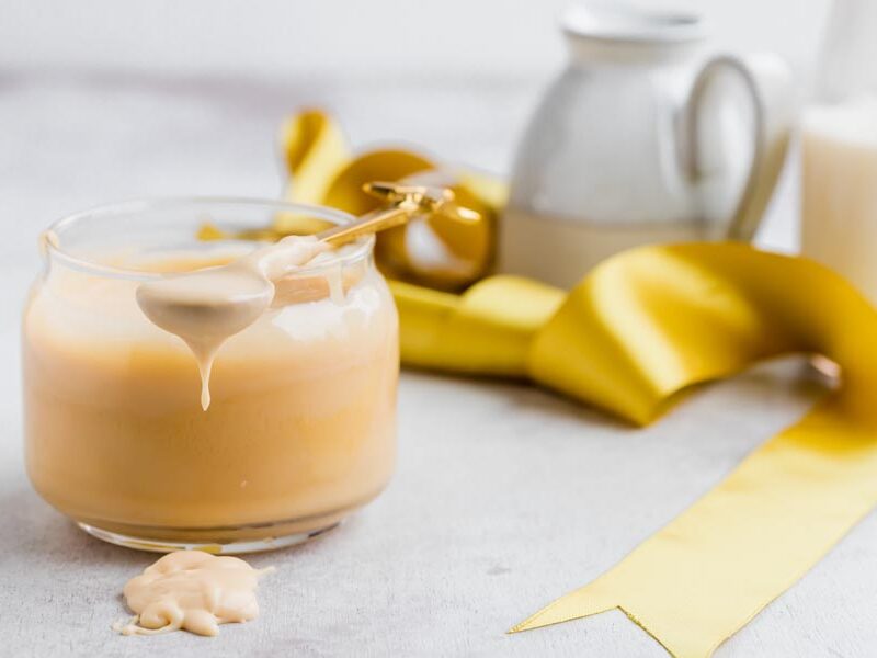 A jar of condensed milk with a spoon resting on top the jar and dripping syrup on the counter. A yellow ribbon is nearby.