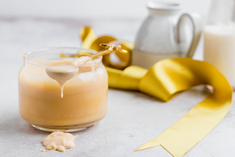 A jar of condensed milk with a spoon resting on top the jar and dripping syrup on the counter. A yellow ribbon is nearby.