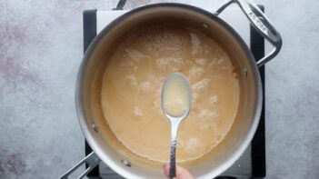 A hand holding a spoon of caramel colored condensed milk over a saucepan.