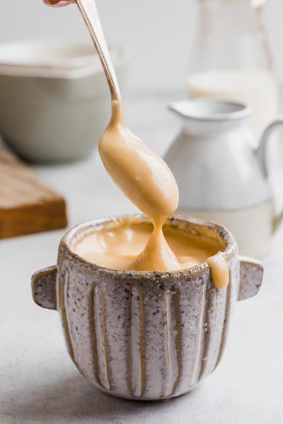 A spoon dripping condensed milk over an rustic cup with a drip of syrup coming down the side.