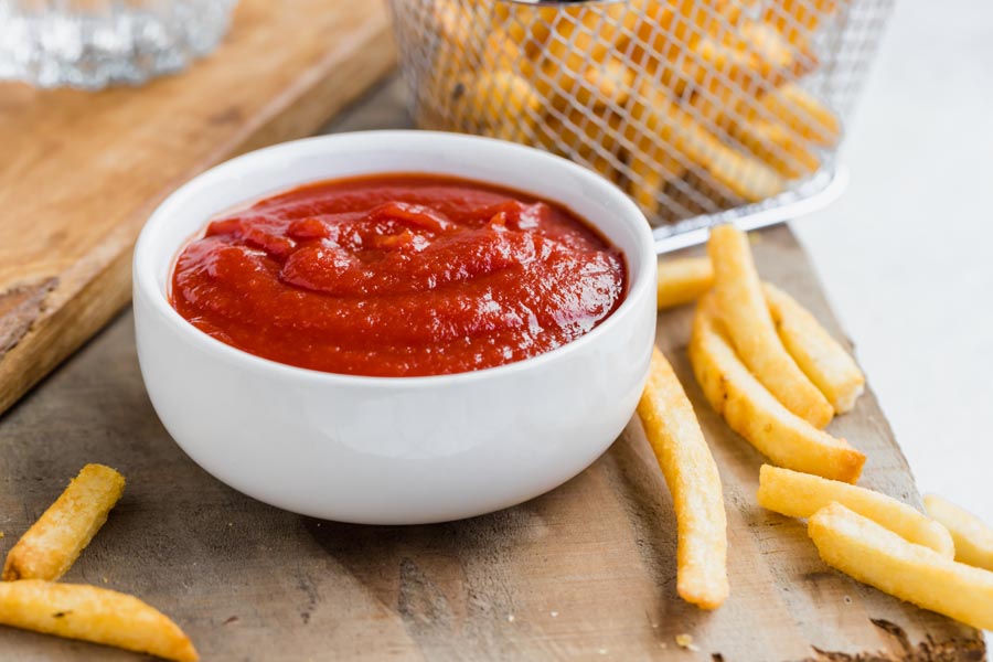 a bowl of thick goopy ketchup sits on a wood board with fries scattered around and a basket of fries behind