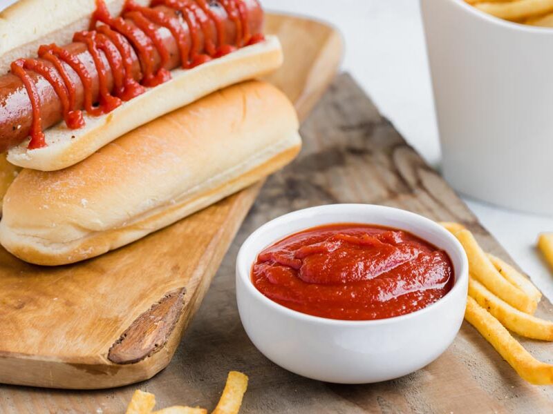 a bowl of ketchup in front of a hot dog in a bun stacked on other buns and fries