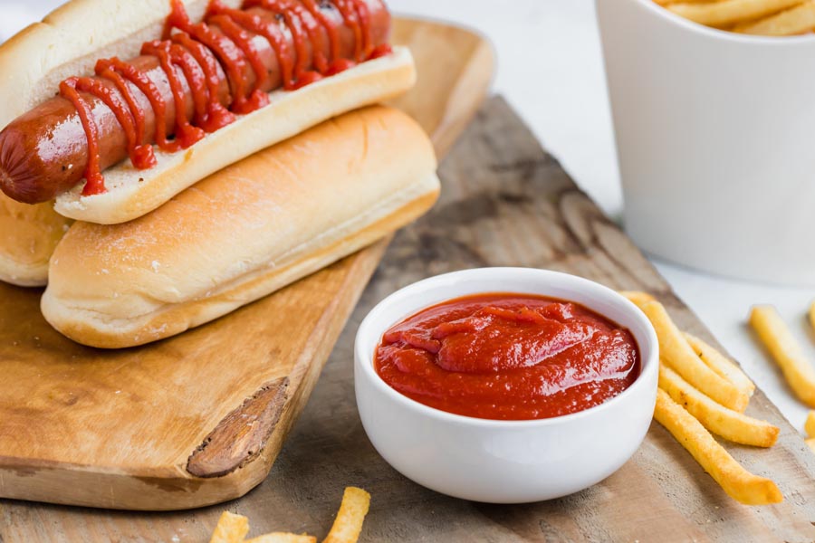 a bowl of ketchup in front of a hot dog in a bun stacked on other buns and fries