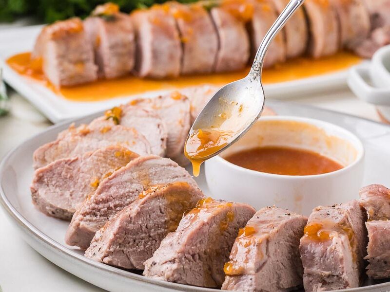 A spoon drizzling sweet orange glaze over roasted pork tenderloin slices beside a bowl of extra sauce.