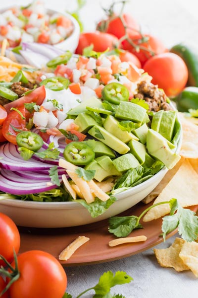 fresh taco salad sits on a terra cotta plate with shredded cheese and avocado falling down