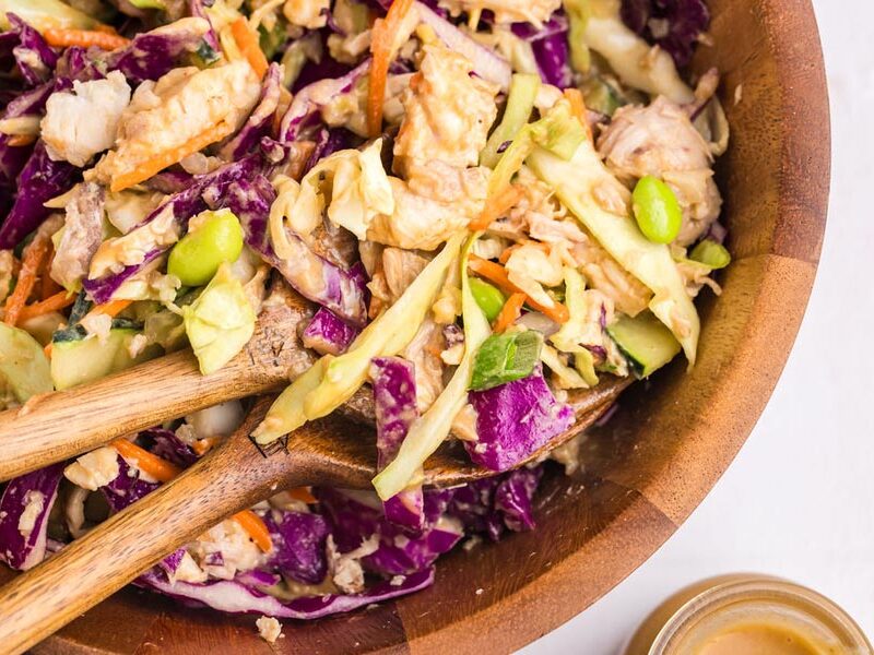 Two serving spoons scooping up helpings of thai chicken salad filled with chicken, cabbage, shelled edamame and carrot.