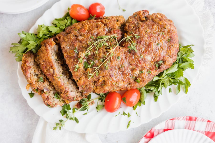 smoked meatloaf on a white platter with vegetables and herbs surrounding