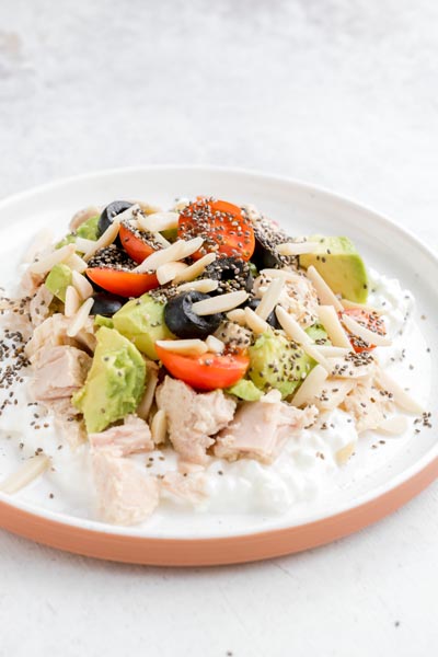 Tuna and cottage cheese bowl topped with avocado, tomatoes, olives, and almonds served on a white plate.