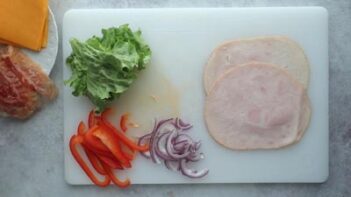 Two slices of turkey meat on a cutting board with sliced bell pepper, onion and lettuce.