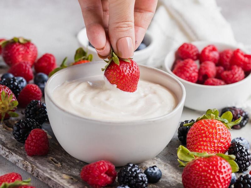 A hand dipping a strawberry into a fruit dip bowl with berries on a fruit tray.