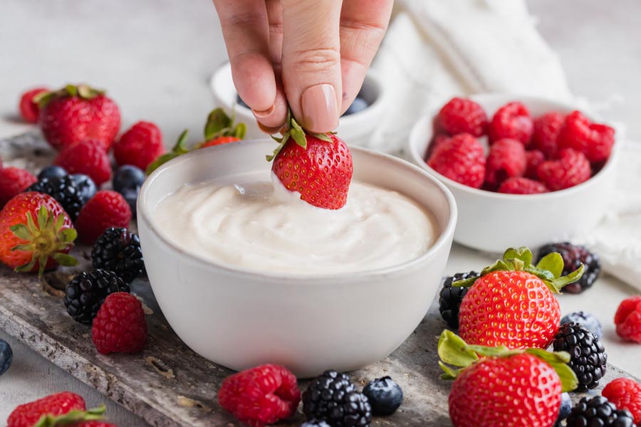 A hand dipping a strawberry into a fruit dip bowl with berries on a fruit tray.