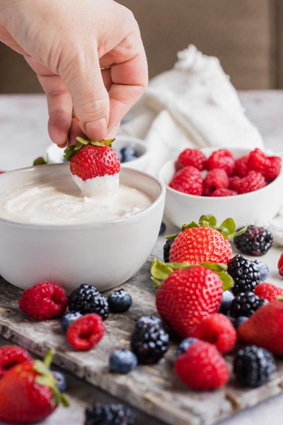 Dipping a fresh strawberry into a dip with a bowl of raspberries to the sides and fresh berries scattered around.